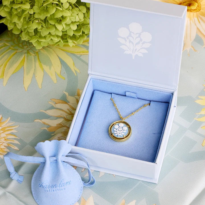 A gold pendant necklace with a white enamel face and clear stones, displayed inside an open blue jewelry box with a light blue velvet lining. A small light blue drawstring pouch with the brand name 'Charm Lane' sits beside the box on a floral surface.