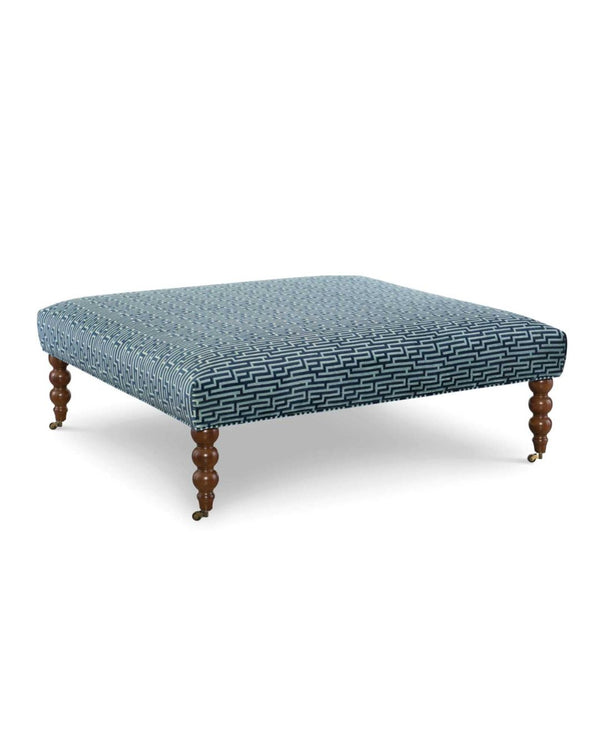 A square ottoman with blue upholstered fabric and wooden bobbin legs with castors.