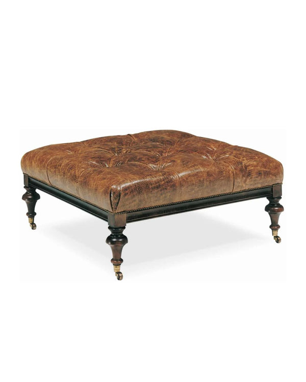 A square brown leather ottoman with tufted upholstery, featuring turned legs and casters.